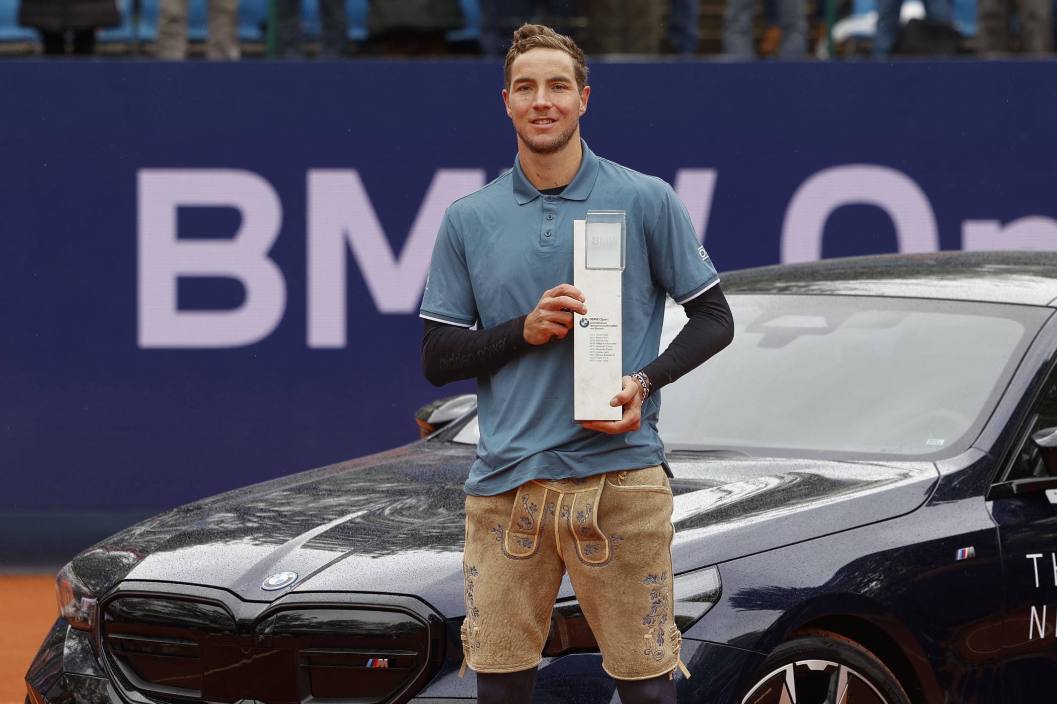 Jan-Lennard Struff posiert in Lederhosen mit einem Pokal vor einem Auto.