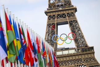 Der Eiffelturm. Oberhalb der ersten Plattform sind die Olympischen Ringe angebracht. Vor dem Turm eine Reihe Nationalflaggen teilnehmender Länder