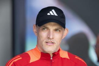 Thomas Tuchel in rotem Trainingsanzug mit orangenen Streifen und schwarzer Adidas-Kappe. Er schaut nach vorne, im Hintergrund ist eine verschwommene Umgebung zu sehen.